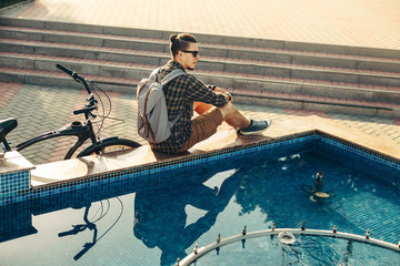 Wall Mural - Young Man Cyclist Sitting Near Fountain Next To Bicycle In Summer Park Daily Lifestyle Urban Resting Concept