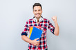 © deagreez - Cheerful young brunet nerdy student with stubble is standing with colorful books on pure background in casual checkered outfit, whitemodern big headphones. He is gesturing victory, he passed exams