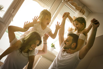 Wall Mural - Family spending free time at home. Daughters sitting on the shoulders of their parents. Fun on bed. Girls sitting on parents shoulders.