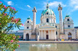 Facade of Karlskirche (St. Charles's Church) against blue sky, white clouds and a pond with water splashes on a hot summer day. Green tree branch with pink flowers on the front. Vienna, Austria