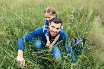 Cheerful girl embracing her father who is walking with her on green summer field, being glad to spend time together. Handsome man with his daughter resting outdoors, admiring beautiful nature