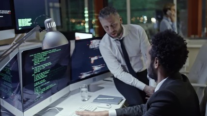 African programmer writing code on computer and talking with his colleague while working in the office at night; businesswoman talking on the phone in background
