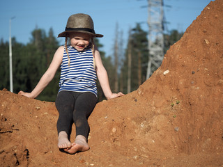 the child is sitting on a mountain of sand. blue sky. a little girl, wearing a hat and barefoot. jou