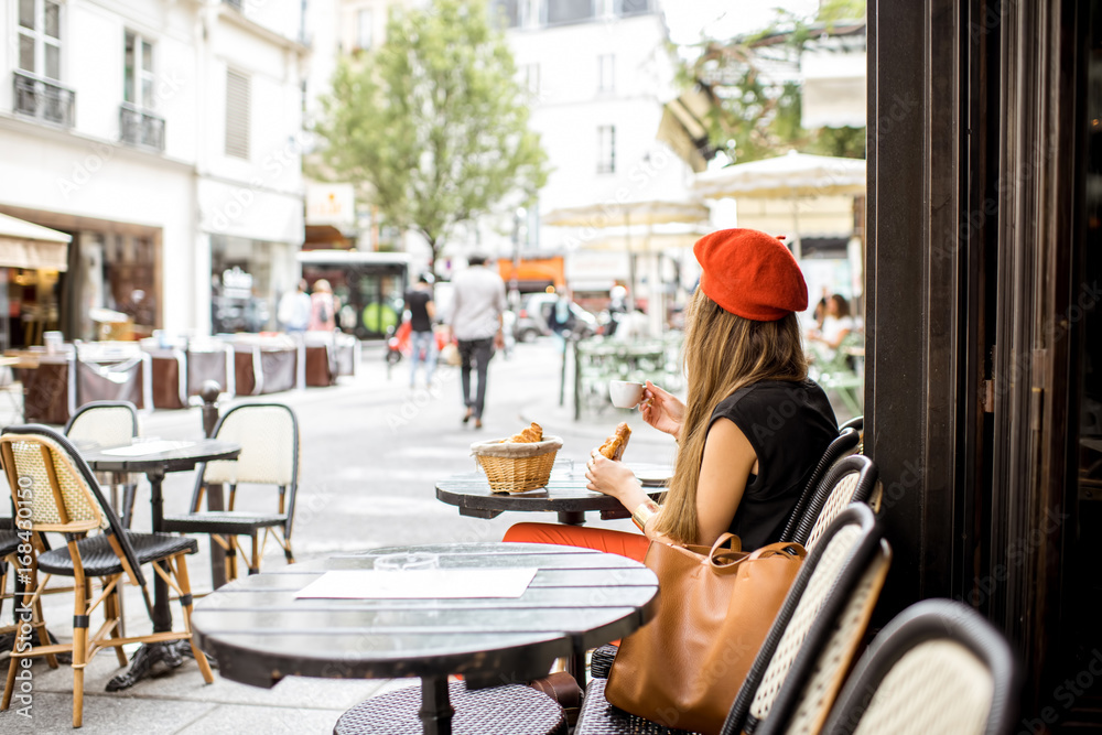 Young stylish woman in red beret having a french breakfast with coffee and croissant sitting oudoors at the cafe terrace - obrazy, fototapety, plakaty 