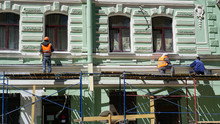 Three Men Are Restoring A Birch Facade Of An Old Building. Men In Red Color Clothes Are Standing On Scaffolding.