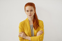Young Caucasian Woman With Red Hair Tied In Pony Tail, Having Appealing Appearance With Freckles, Looking With Serious Expression While Keeping Her Hands Crossed, Isolated Over White Background
