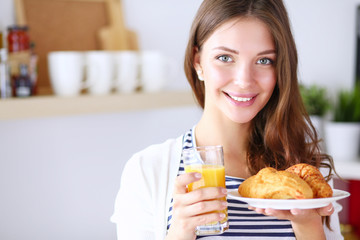 Young woman with glass of juice and cakes standing in kitchen