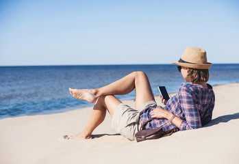 Wall Mural - Young woman lying on a beach with smart phone. Technology and travel concept. People using mobile devices to stay connected from remote parts of the world