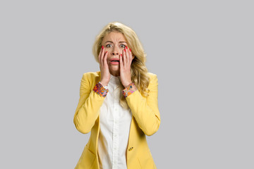 Portrait of horrified young woman. Desperate young blond woman touching her face looking confused and terrified, grey background.