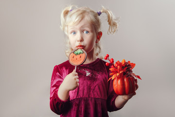 Portrait of adorable funny white Caucasian girl with pigtails dressed for Halloween. Child playing, having fun, making faces in studio for autumn fall seasonal holiday. Kid eating chocolate lollipop