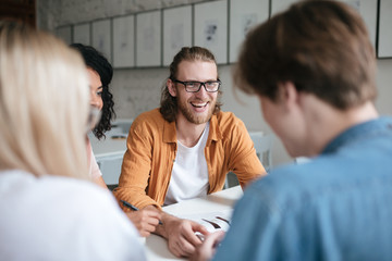 Wall Mural - Portrait of young smiling man with blond hair and beard happily looking at friends. Joyful boy in glasses sitting in office and working with friends