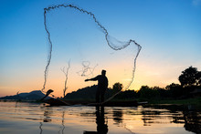 Asian Fisherman On Wooden Boat Casting A Net For Catching Freshwater Fish In Nature River In The Early Morning Before Sunrise.