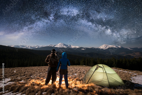 Romantic Couple Tourists Enjoying In The Camping At Night Holding Hands Standing Near Green Tent Under Beautiful Night Sky Full Of Stars And Milky Way On The Background Snow Covered Mountains Buy