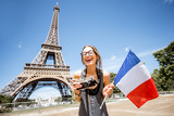 Fototapeta Paryż - Young and happy woman tourist with french flag and photo camera in front of the Eiffel tower in Paris