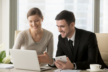 Smiling happy businessman and businesswoman sitting at workplace, using laptop and digital tablet, smart devices for easy business management, enterprise software and apps, integrated tech solutions