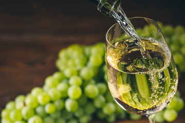 Pouring white wine into a glass with a bunch of green grapes against wooden background