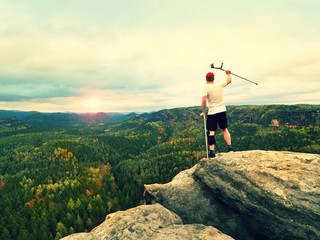Hiker with knee joint bandage and forearm poles. Hiker on cliff makes triumph gesture.