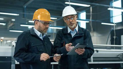 two engineers in hardhats discuss information on a tablet computer while standing in a factory.