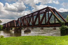 Old Rusted Railroad Bridge Crossing A River With Green Grass, Mountains In The Distance And A Cloudy Blue Sky Overhead.