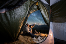 View From Inside A Tent On The Two Tourists Have A Rest In The Camping In The Mountains At Night. Men Sitting Near Campfire. One Guy Is Pointing At The Beautiful Night Sky Full Of Stars And Milky Way