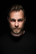 © _robbie_ - Portrait of a bearded man with an intense look standing in a dark room against a black background.