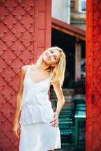 Blonde Fasion Stylish Woman In Whiti Dress Posing Over Old Red Door In Summer Morning.