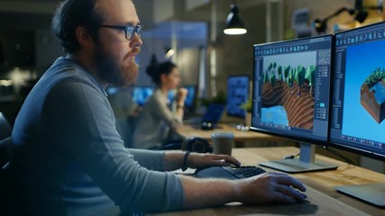 Poster - Creative Male Game Developer Works with Graphics for the New Level Design, He Sits at His Wooden Table Working on Two Display Personal Computer. Other People Work in This Creative Studio.