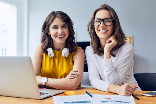 Couple of gorgeous young businesswomen smiling and staring straight at camera while working in office