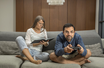 Wall Mural - couple on a sofa, man is playing video games while woman reading a book