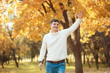 Young handsome man in sweater and jeans staying in autumn orange park smiling and waving to someone. Great weather for walk and spending time with friends. Emotions, people, relations, business.