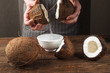 © golubovy - Fresh coconut water pouring into white bowl on wooden table. Preparing of beauty products, cosmetic oils and body lotions from organic ingredients of tropical fruits