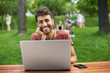 © Cookie Studio - Stylish bearded hipster in red t-shirt sitting on bench at the park, working outdoors on his notebook. Caucasian male student enjoying summer day.
