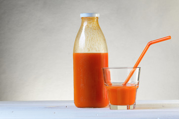 A bottle of carrot juice and a glass of carrot juice with a straw on white wood table. Still life of a healthy eating concept