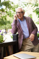 Wall Mural - Portrait of modern senior man speaking by phone outdoors in cafe, leaning on flowerbed