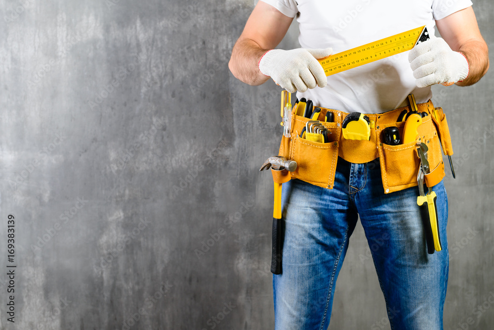 unidentified handyman standing with a tool belt with construction tools and holding roulette against grey background with copyspace. DIY tools and manual work concept - obrazy, fototapety, plakaty 
