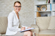 Portrait of beautiful female psychologist wearing glasses posing with clipboard in therapy office smiling to camera