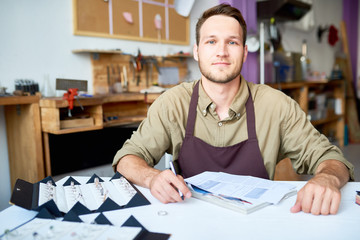 Wall Mural - Portrait of smiling young man sitting at table in jeweler shop looking at camera with documentation and ring  set in front of him