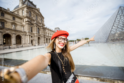 Young woman tourist in red cap making selfie photo in front of the famous Louvre Poster