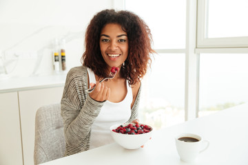 Wall Mural - Smiling african woman eating by the table in kitchen