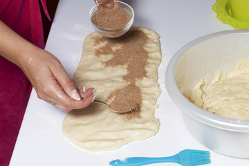 A woman is cooking buns with cinnamon. She rolls the stuffing onto the rolled dough. Mixed sugar and cinnamon.