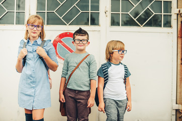 Group of three funny kids wearing backpacks walking back to school. Girl and boys wearing eyeglasses posing outdoors