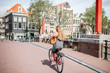 Young beautiful woman riding a bicycle with bag and bouquet of tulips on the bridge over the water channel in Amsterdam old city