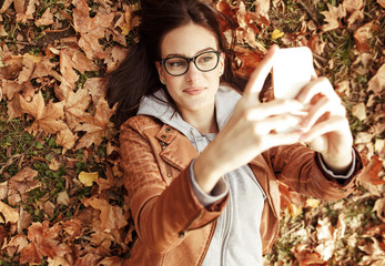 Portrait of young urban style woman lying down at park and taking image with smart phone.Autumn season.