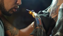 Tattooist Does Tattoo A Client With Machine Close Up. Process Getting Tattoo In A Parlor. Body Tattooing Process. Man With Dreadlocks Working On Female Body Part Under Lamp. Slow Motion.