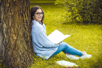 A beautiful girl in glasses, reading a book in nature. A student near the university, resting in the park.