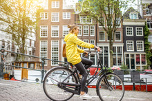 Young Woman In Yellow Raincoat With Bag And Flowers Riding A Bicycle In Amsterdam City