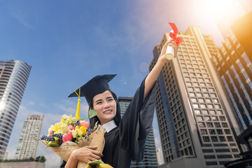 Beautiful woman graduating holding  diploma with pride and smiling in an academic gown.Graduate woman student wearing graduation hat and gown ,background is modern education building in university.