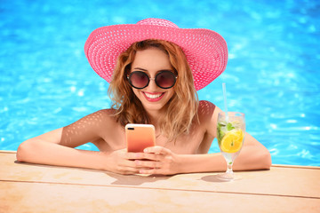 Wall Mural - Beautiful young woman with glass of lemonade and mobile phone in swimming pool