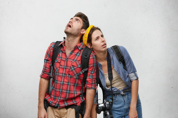 Wall Mural - Studio shot of young man and woman travelers in sensible clothes carrying heavy backpacks leaning on each other, feeling exhausted and thirsty while hiking. Travel, peolple and relationships concept