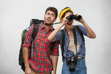 Wall Mural - Young European couple on safari trip: happy woman using binoculars while examining area, pulling strap making her unshaven boyfriend feel uncomfortable. Tourism, travel, relationship and adventure